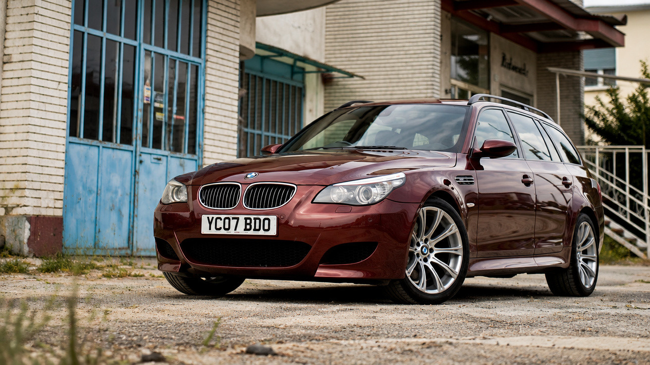 Maroon BMW estate car parked on a gravel surface in front of a building with blue doors.